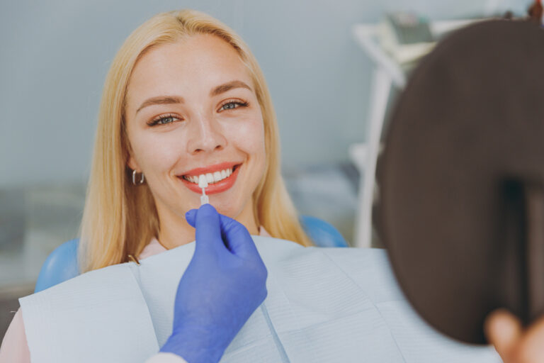 Young smiling calm happy woman choose veneers enamel color compare teeth shade with stomatologist look at result mirror sit at dentist office chair indoor modern cabinet. Enamel whitening treatment.