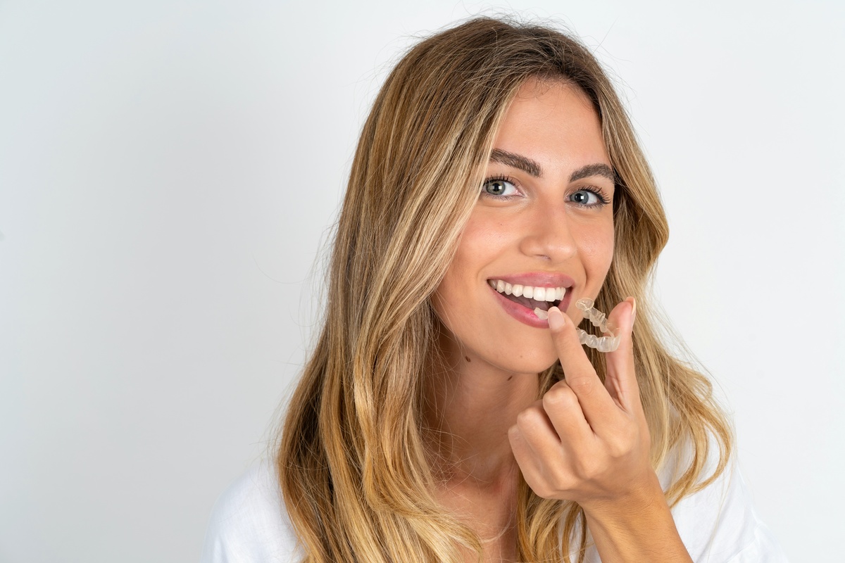 young blonde woman wearing white shirt over white studio background holding an invisible aligner ready to use it. Dental healthcare and confidence concept.